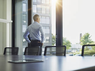 Mature businessman with hands on hips looking out through window - RORF02896