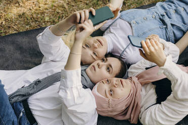 Friends using smart phones lying on picnic blanket at park - KASF00013