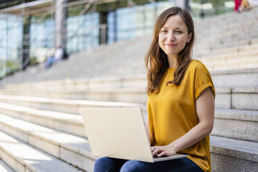 Lächelnde Geschäftsfrau mit Laptop auf Stufen sitzend - DIGF18242