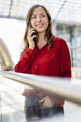 Lächelnde Frau mit Hand in der Tasche, die mit ihrem Smartphone telefoniert und am Geländer steht - DIGF18260