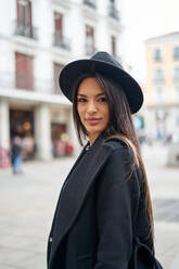 Attractive female in coat and stylish hat looking at camera while standing on street with buildings in city - ADSF35741