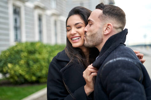 Positiver Freund, der die Wange seiner lächelnden Freundin in Oberbekleidung küsst, während er während eines romantischen Dates in der Stadt auf der Straße in der Nähe eines Gebäudes steht - ADSF35743