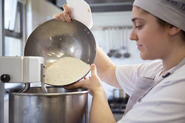 Seitenansicht einer Bäckerin in Uniform, die bei der Herstellung von Teig in einer Bäckerei Mehl in die Schüssel eines Mixers gießt - ADSF35744