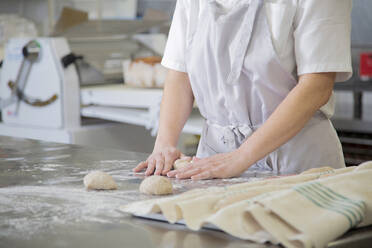 Anonymer weiblicher Koch in Uniform, der Mehl auf einem Tisch mit Kugeln aus rohem Teig verteilt, während er in einer Bäckerei mit verschiedenen Geräten arbeitet - ADSF35746