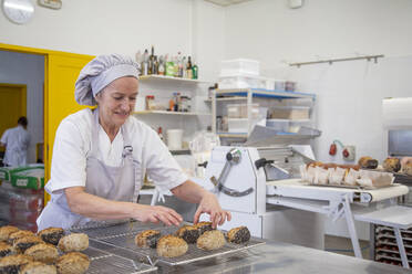 Fokussierte Bäckerin in Uniform mit gebackenem Brot auf einem Metalltisch in einer modernen Bäckereischule - ADSF35747