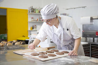 Konzentrierte Bäckerin in Uniform bereitet Teig für Kekse auf einem Metalltisch vor, während sie in einer leichten Backstube mit verschiedenen Geräten arbeitet - ADSF35748