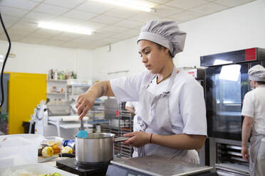 Weibliche Köchin in Uniform, die Zutaten in einem Topf umrührt, während sie auf dem Herd Sirup zubereitet, in einer Berufsschule für Leichtbäckerei - ADSF35750