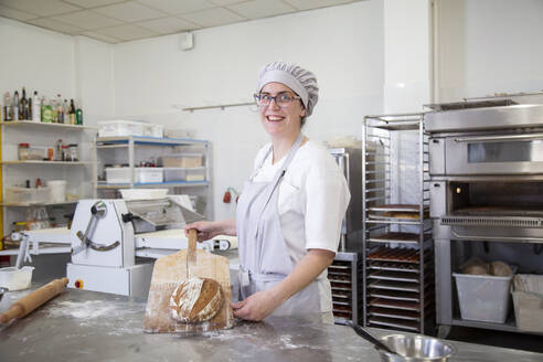 Cheerful female baker in uniform showing tasty baked bread loaf and looking at camera while standing in bakery with various equipment - ADSF35751