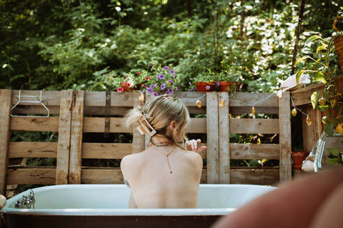 Rückansicht einer gesichtslosen, nachdenklichen Frau, die in einer Badewanne in der Nähe eines Holzzauns auf einer Terrasse sitzt, während sie sich an einem Sommertag um ihre Haut kümmert - ADSF35779