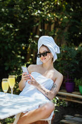 Zufriedene Frau, eingewickelt in Handtücher, die eine SMS auf ihrem Handy schreibt, während sie an einem Tisch mit einem Glas Saft auf der Terrasse eines Gebäudes sitzt - ADSF35780