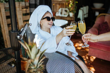 Woman wrapped in towels clinking glasses of fresh juice with crop anonymous person while sitting on terrace on summer day - ADSF35784