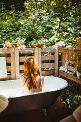 Rückansicht einer gesichtslosen, nachdenklichen Rothaarigen, die in der Badewanne in der Nähe des Holzzauns auf der Terrasse sitzt, während der Hautpflege am Sommertag - ADSF35791