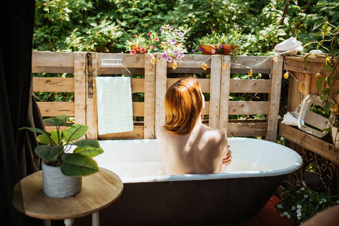 Rückansicht einer gesichtslosen, nachdenklichen Rothaarigen, die in der Badewanne in der Nähe des Holzzauns auf der Terrasse sitzt, während der Hautpflege am Sommertag - ADSF35792