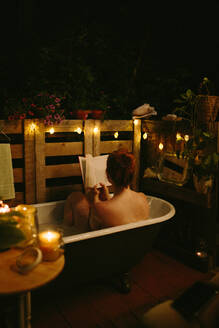 Rückenansicht einer nicht erkennbaren nackten Frau, die ihr Gesicht mit einem Buch bedeckt, während sie in einer Badewanne sitzt, die in einem Innenhof mit einer leuchtenden Girlande in der Abendzeit aufgestellt ist - ADSF35798