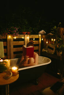 Unerkennbare nackte Frau, die ihr Gesicht mit einem Buch bedeckt, während sie in einer Badewanne sitzt, die im Innenhof mit einer leuchtenden Girlande in der Abendzeit aufgestellt ist - ADSF35799