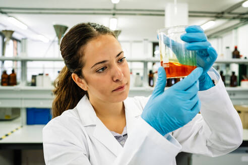 Fokussierte Wissenschaftlerin mit Schutzbrille und Latexhandschuhen, die einen Kolben mit einer hellen chemischen Flüssigkeit beobachtet, während sie in einem hellen Labor forscht - ADSF35800