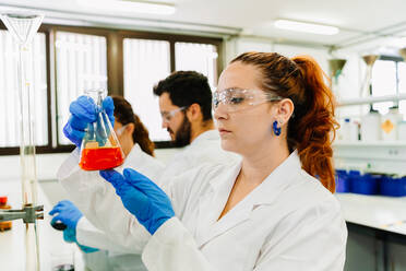 Fokussierte Wissenschaftlerin mit Schutzbrille und Latexhandschuhen, die einen Kolben mit einer hellen chemischen Flüssigkeit beobachtet, während sie in einem hellen Labor forscht - ADSF35809