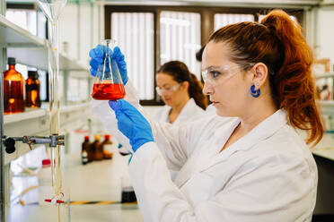 Focused female scientist in protective glasses and latex gloves observing flask with bright chemical liquid while doing research in light laboratory - ADSF35811