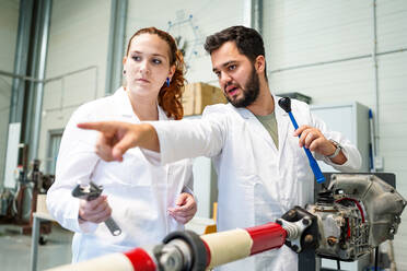 Wissenschaftler in medizinischer Uniform zeigt von einer weiblichen Kollegin weg, die mit einem Schraubenschlüssel in der Nähe eines speziellen Geräts mit Metallrohr während eines Reparaturvorgangs in einem modernen Labor steht - ADSF35813