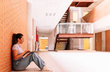 Ganzkörper-Seitenansicht einer begeisterten Studentin in Freizeitkleidung, die im Internet auf einem Netbook surft, während sie auf dem Boden in der Nähe einer Backsteinmauer sitzt - ADSF35818