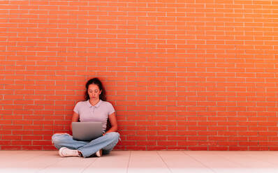 Full body of focused female student in casual wear surfing internet on netbook while sitting on floor near brick wall - ADSF35819