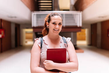 Optimistische Studentin mit Notizblock in den Händen, die lächelnd in die Kamera schaut, während sie in der Aula einer modernen hellen Universität steht - ADSF35820