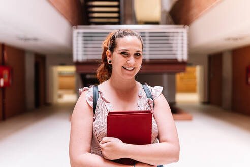 Optimistic female student with notepad in hands looking at camera with smile while standing in hall of modern light university - ADSF35820