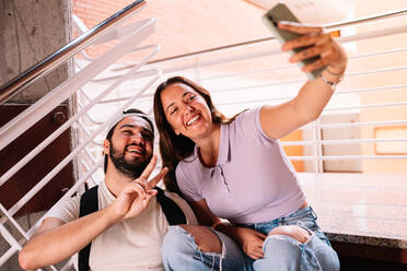 Ein paar begeisterte Studenten in Freizeitkleidung nehmen ein Selbstporträt mit ihrem Handy auf, während sie zusammen auf einer Treppe im Flur der Universität sitzen - ADSF35822