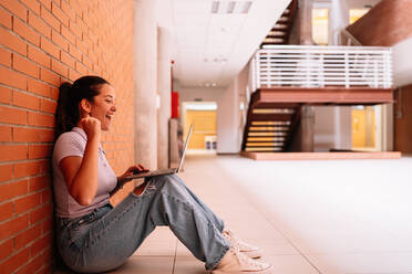 Seitenansicht des gesamten Körpers einer aufgeregten Studentin in Freizeitkleidung, die im Internet auf einem Netbook surft, während sie auf dem Boden in der Nähe einer Backsteinmauer sitzt - ADSF35823