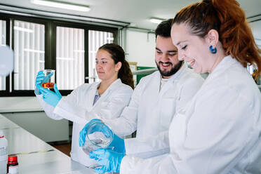 Gruppe positiver Kollegen in weißen Uniformen, die eine chemische Flüssigkeit vorbereiten, während sie an einem Tisch während der Arbeit in einem modernen Labor forschen - ADSF35826