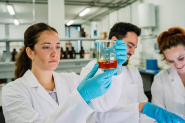 Focused female scientist in protective glasses and latex gloves observing flask with bright chemical liquid while doing research in light laboratory - ADSF35827