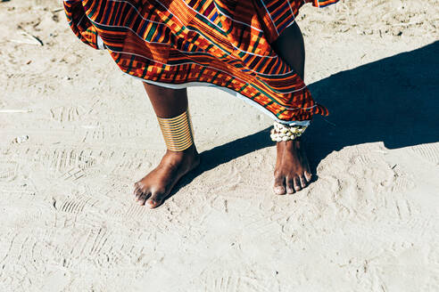 Nicht erkennbare ethnische Frau in farbenfrohem handgewebtem Kente trägt Kayan-Ringe am Knöchel, während sie am Sandstrand steht - ADSF35835