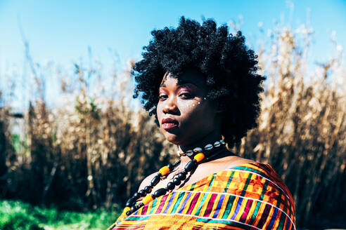 Seriöses afrikanisches Modell mit Afrofrisur, das bunte handgewebte Kente trägt und in die Kamera schaut, während es auf einem Feld an einem hellen Sommertag steht - ADSF35836