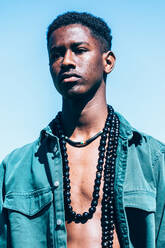Junger schwarzer Mann in stilvollem Jeans-Outfit mit traditioneller Halskette schaut weg, während er auf grünem Gras gegen blauen Himmel in ländlichem Gelände steht - ADSF35851