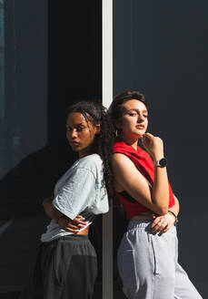 Ernsthafte multirassische Freundinnen in Freizeitkleidung schauen in die Kamera, während sie in der Nähe einer grauen Gebäudewand auf einer sonnigen Straße stehen - ADSF35862