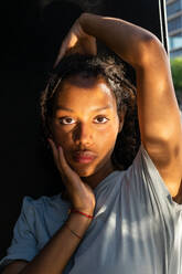Junge Afroamerikanerin mit langen, dunklen, lockigen Haaren in weißem T-Shirt, die nachdenklich in die Kamera blickt, in der Nähe eines Fensters im Sonnenlicht - ADSF35876