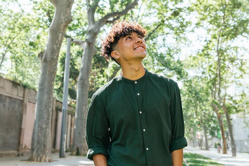 Young male looking away while standing on grassy pathway in city park on bright summer day - ADSF35879