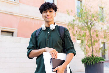 Niedriger Winkel eines lächelnden männlichen Studenten mit Netbook und Kopfhörern, der in die Kamera schaut, während er auf einer Straße in der Nähe eines hohen Gebäudes steht und den sonnigen Sommertag genießt - ADSF35881