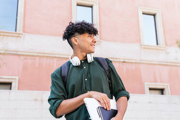 Niedriger Winkel eines lächelnden männlichen Studenten mit Netbook und Kopfhörern, der wegschaut, während er auf einer Straße in der Nähe eines hohen Gebäudes steht und den sonnigen Sommertag genießt - ADSF35882