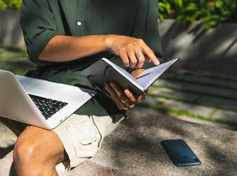 Unbekannter männlicher Student sitzt auf einer Steinbank im Stadtpark und liest Notizen in einem Notizblock, während er mit einem modernen Netbook an einer Aufgabe arbeitet - ADSF35885