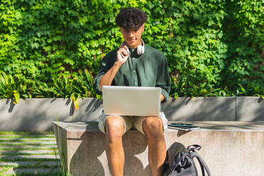 Lächelnder junger Mann in Freizeitkleidung sitzt auf einer Steinbank in einem grünen Park und arbeitet an einem Fernprojekt mit einem modernen Netbook auf der Straße - ADSF35886