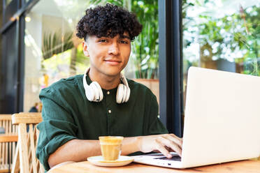 Ethnischer männlicher Freiberufler mit lockigem Haar trinkt Kaffee, während er in einem Café an einem modernen Netbook arbeitet - ADSF35887