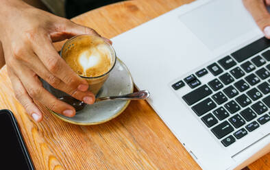 Anonymous ethnic male freelancer drinking coffee while working remotely on modern netbook in cafe - ADSF35888