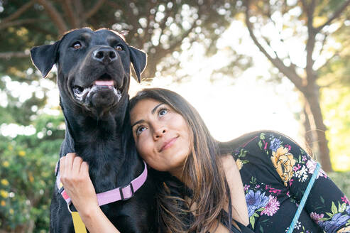 Fröhliche Besitzerin mit langen Haaren, die ihren treuen Labrador Retriever-Hund mit schwarzem Fell umarmt und streichelt, während sie an einem Sommertag im Park in der Nähe eines Baumstamms ruht - ADSF35907