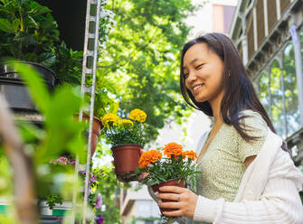 Positive asiatische Frau mit blühenden Ringelblumen in Töpfen, die wegschaut, während sie in der Nähe von Regalen mit verschiedenen Blumen auf dem Markt steht - ADSF35909