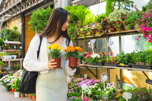 Positive Asian female with blooming marigolds in pots looking away while standing near shelves with assorted flowers in market - ADSF35910