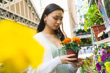 Seitenansicht Asiatische Frau mit blühenden Ringelblumen in Töpfen, die wegschaut, während sie in der Nähe von Regalen mit verschiedenen Blumen auf dem Markt steht - ADSF35911