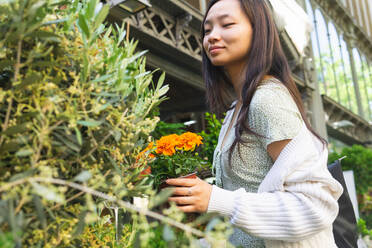 Seitenansicht einer fröhlichen asiatischen Frau, die bunte Topfblumen auswählt, während sie in einem hellen Blumenmarkt mit einer Fülle von Pflanzen steht - ADSF35913