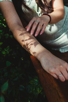 Crop anonymous female with shadow of sprig on arm sitting on bench in park on sunny summer day in city - ADSF35914