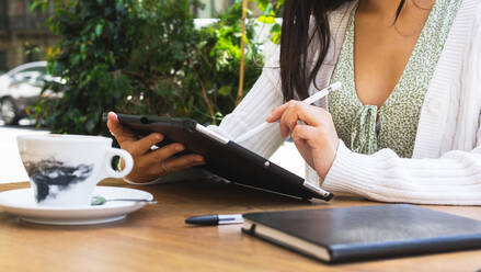 Cropped unerkennbar weiblichen Freiberufler Schreiben auf Tablet mit Stift beim Sitzen auf der Terrasse der Cafeteria am Sommertag in der Stadt - ADSF35918